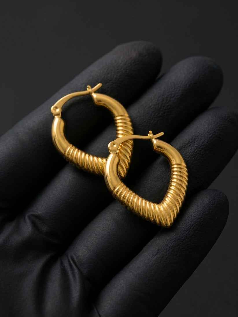 Gold heart hoop earrings held by a black gloved hand against a dark background