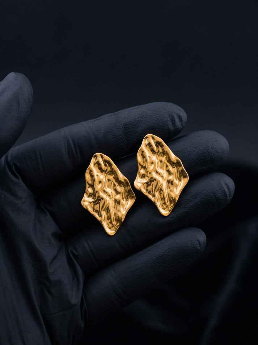 Gold irregular earrings held in a black gloved hand against a dark background