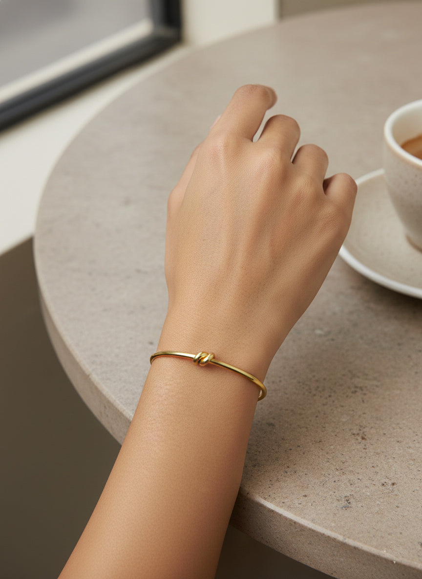 Hand wearing a stainless steel gold bracelet with knot design on a table with a cup of coffee
